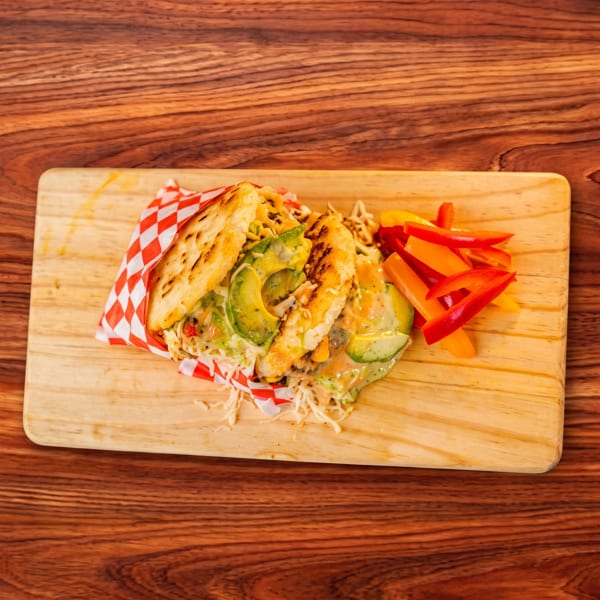 Arepa with chicken, avocado, and shredded cabbage, served with bell pepper strips on a wooden board.