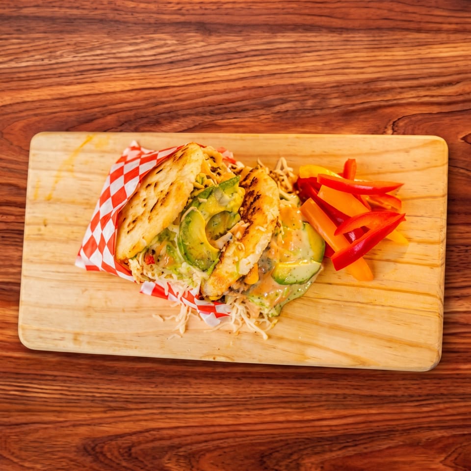 Arepa with chicken, avocado, and shredded cabbage, served with bell pepper strips on a wooden board.