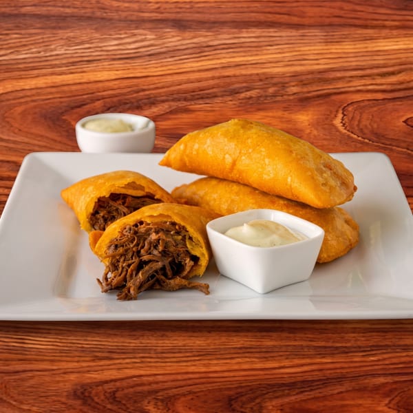 Golden-brown empanadas, some open revealing shredded meat filling, with dipping sauces on a white plate.